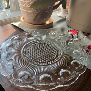 Könnte beinhalten: Eine klare Glasservierplatte mit einer erhabenen, strukturierten Mitte und einem gewellten Rand. Die Platte hat ein dekoratives Muster aus kleinen Kreisen am Rand.