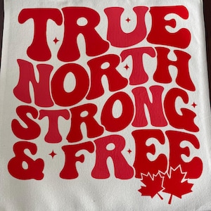 May include: A white canvas tote bag featuring the words "TRUE NORTH STRONG & FREE" in red and pink lettering. The text is in a retro font style. Two red maple leaves are at the bottom of the bag.