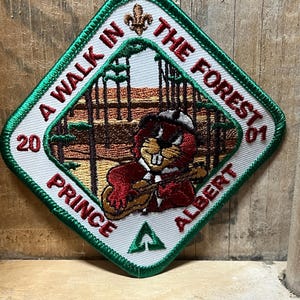 May include: Embroidered patch with a green border and the text "A Walk in the Forest" and "Prince Albert". The patch features a cartoon beaver playing a guitar in a forest scene. The patch is diamond-shaped.