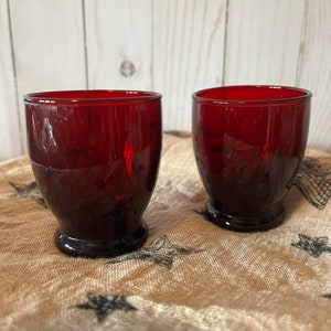 May include: Two ruby red glass tumblers with a rounded base. The glasses are sitting on a brown fabric with a star pattern.