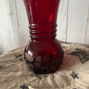 May include: A deep red glass vase with a round body and a flared top. The vase has a textured design on the bottom.