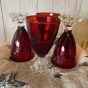 May include: Three red glass goblets with clear, beaded stems. The goblets are sitting on a white fabric with a black star pattern.