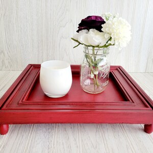 以下が含まれることがあります： 4本の脚と縁が盛り上がった赤い木製のトレイ。トレイには白いろうそくと、白と紫の花が入ったガラスの瓶が置かれている。