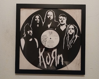 May include: Framed black and white artwork featuring the band Korn. The artwork is a vinyl record with a silhouette of the band members and the band's logo. The frame is black and the background is a light color.