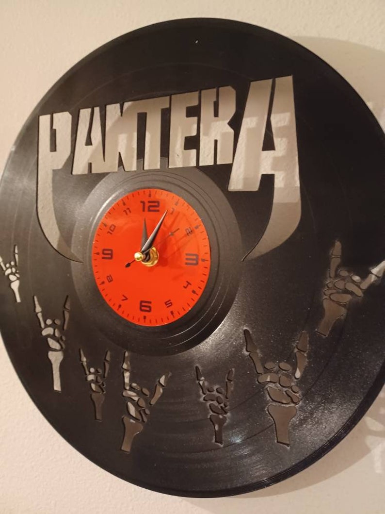 Może przedstawiać: Czarny zegar winylowy z napisem "PANTERA" w srebrnych literach. Tarcza zegara jest czerwona z czarnymi cyframi i wskaz&oacute;wkami. Płyta ma wz&oacute;r szkieletowych dłoni wykonujących gest rock and roll.