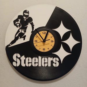 May include: Black and white vinyl record clock with a silhouette of a football player and the word "Steelers" in white. The clock face is yellow with black numbers.