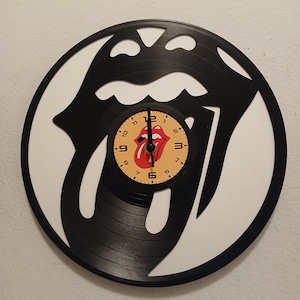 May include: A black vinyl record clock with a white border. The clock features the iconic Rolling Stones tongue and lips logo. The clock face is yellow with black numbers and hands. The Rolling Stones logo is red.