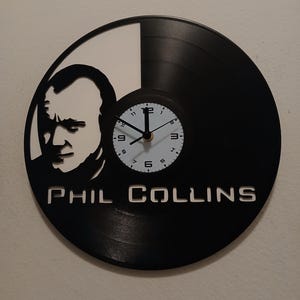 May include: A black vinyl record clock featuring a silhouette of Phil Collins with the text "PHIL COLLINS" printed below. The clock face is white with black numbers and hands.
