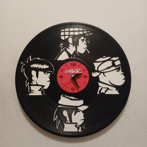 May include: Black vinyl record clock featuring white silhouettes of the Gorillaz band members around a red clock face. The clock face has the word "Gorillaz" and the numbers 1 through 12. The clock hands are red.