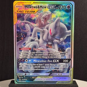 Puede incluir: Una carta coleccionable Pokémon holográfica con Mewtwo & Mew GX. La carta tiene un fondo colorido con el texto "Mewtwo & Mew GX HP270" en la parte superior. La carta se muestra en un soporte negro.