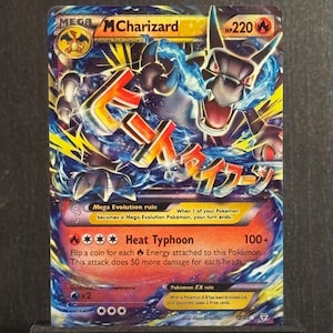 May include: A collectible Mega MCharizard Pokémon card. The card features a vibrant illustration of a dragon-like creature with blue and orange accents. The card has the text "Heat Typhoon" and "HP220". The card is displayed on a black stand.