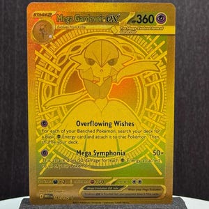 May include: A gold-colored Mega Gardevoir EX trading card. The card features a detailed illustration of Gardevoir with the text "Mega Gardevoir EX" and "HP 360". The card is displayed on a black stand.