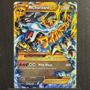 May include: A Pokémon trading card featuring Mega MCharizard EX. The card has a gold and blue color scheme with Japanese text and a dragon illustration. The card is displayed on a black stand.