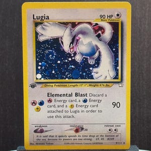 May include: A Pokémon trading card featuring Lugia, a Diving Pokémon. The card has a yellow border and displays a white and blue illustration of Lugia. Text includes "Elemental Blast" and "90 HP". The card is 5.21m long and weighs 215.9 kg.