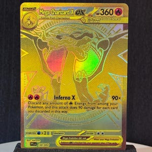 Puede incluir: Una tarjeta Pokémon Mega Charizard X EX de lámina dorada, exhibida en un soporte negro. La tarjeta presenta una ilustración detallada de Charizard con acentos rojos, verdes y amarillos. El texto incluye "Inferno X" y "HP 360".