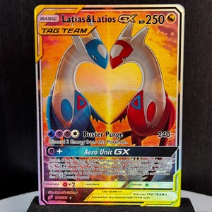 May include: A Pokémon trading card featuring Latias & Latios GX, a Tag Team. The card has a gold background with two stylized creatures in blue and red, forming a heart shape. The card is displayed on a black stand.