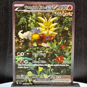 May include: A Gouging Fire EX Pokémon card with a vibrant illustration of a fiery creature in a jungle setting. The card features text including "Heat Blast" and "Blaze Blitz", with a HP of 230. The card is displayed on a black stand.
