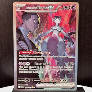 May include: A Pokémon trading card with a holographic design, showcasing Mewtwo EX and a Team Rocket character. The card has a red and purple backdrop, with text including "Team Rocket's Mewtwo EX" and "Erasure Ball". It is presented on a black stand.
