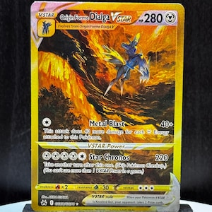 May include: A Pokémon trading card featuring Origin Forme Dialga VSTAR. The card has a gold background with a fiery design and a blue and black creature. Text includes "Metal Blast" and "Star Chronos".
