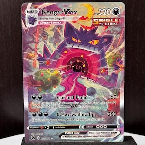 May include: A Gengar VMAX Pokémon card featuring a purple Gigantamax Gengar with a red and pink background. The card displays the text "Fear and Panic" and "G-Max Swallow Up". The card is displayed on a black stand.