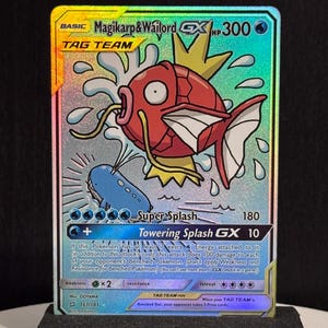 May include: A holographic Pokémon trading card featuring Magikarp & Wailord GX. The card shows a red Magikarp and a blue Wailord with the text "Super Splash" and "Towering Splash GX". The card is displayed on a black stand.