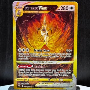 以下が含まれることがあります： 金と赤のアセウスVSTARポケモンカード。カードには「Trinity Nova」と「Starbirth」の文字が入った白と金のキャラクターが描かれています。カードは黒いスタンドに展示されています。
