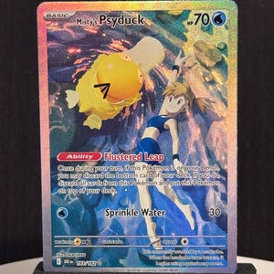 Puede incluir: Una tarjeta coleccionable holográfica con "Misty's Psyduck". La tarjeta muestra un personaje Psyduck amarillo y una mujer con un atuendo azul. El texto incluye "Flustered Leap" y "Sprinkle Water". La tarjeta está apoyada en un soporte negro.