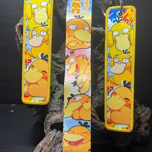 May include: Two rectangular embroidered keychains and one fabric lanyard featuring a repeating pattern of the yellow character Psyduck. The items display various expressions of the character against colourful backgrounds with the text Psyduck.