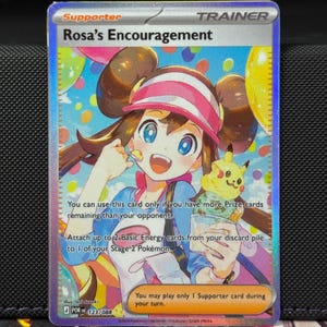 May include: A holographic Pokémon trading card featuring Rosa's Encouragement. The card shows a smiling girl in a pink and white hat, holding a spoon and a dessert with a Pikachu. The card has text that describes its use.
