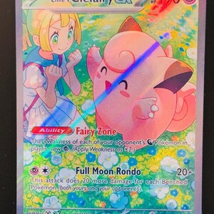 May include: Lillie's Clefairy EX Pokemon card. The card features a pink Clefairy with a girl in a blue and white dress. The card has a light blue background with white flowers. The card's text reads "Fairy Zone" and "Full Moon Rondo".