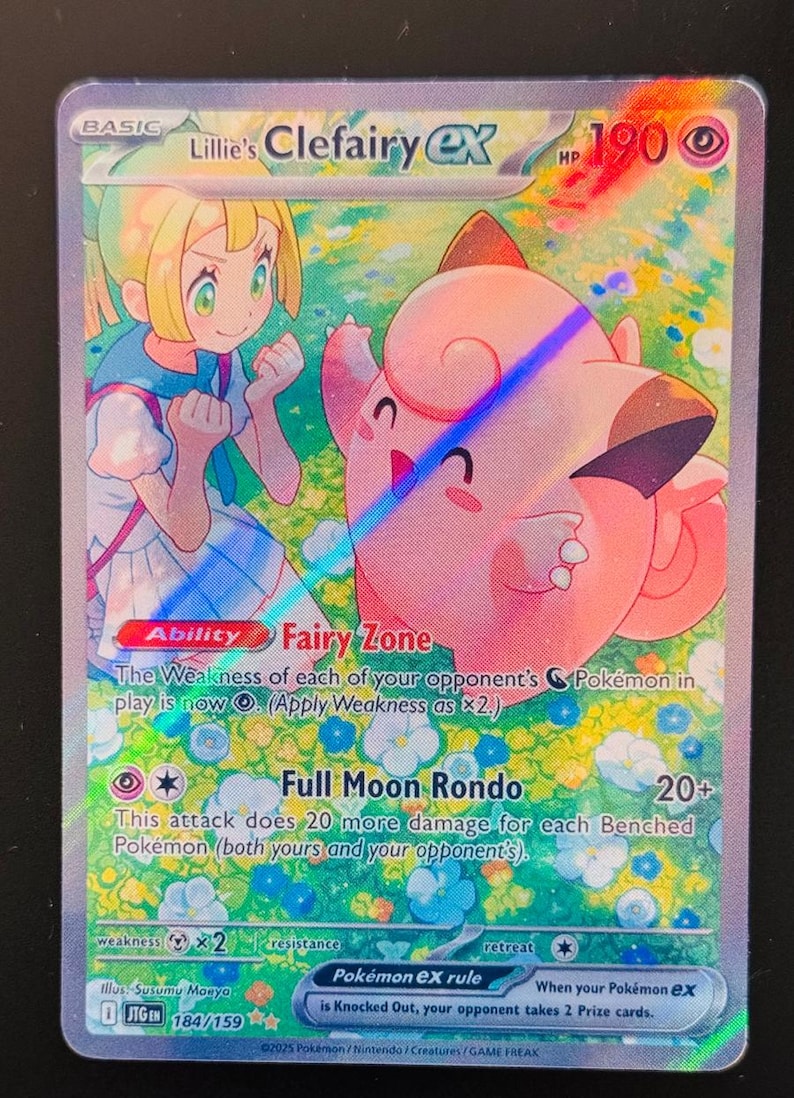 May include: Lillie's Clefairy EX Pokemon card. The card features a pink Clefairy with a girl in a blue and white dress. The card has a light blue background with white flowers. The card's text reads "Fairy Zone" and "Full Moon Rondo".