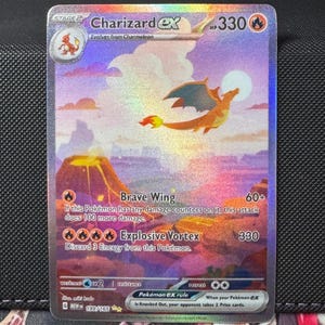 May include: A holographic Charizard EX Pokémon card. The card features a flying orange dragon with blue wings against a colorful, fantasy landscape. Text includes "Brave Wing" and "Explosive Vortex".