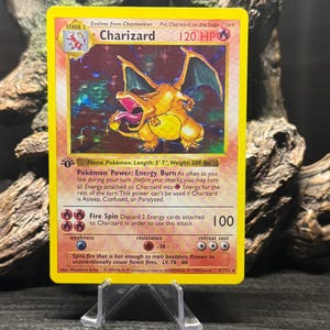 May include: A holographic Charizard trading card featuring a yellow border, 120 HP, and the Flame Pokémon illustration. The card text includes Pokémon Power Energy Burn, Fire Spin attack, and stats of 5' 7" length and 200 lbs. weight.
