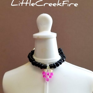 May include: A black beaded necklace with a pink heart charm. The necklace is on a white mannequin.