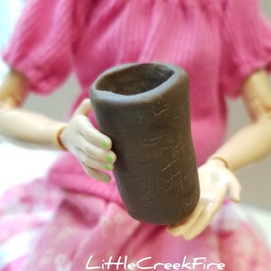 May include: A small, brown, cylindrical ceramic pot with a textured surface. The pot is being held by a person's hands.