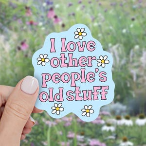 May include: A light blue die-cut sticker with a wavy edge featuring the pink text I love other people's old stuff. The sticker is decorated with five white daisies with yellow centers arranged around the lettering in a playful, casual font.