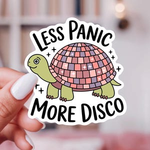 May include: A die-cut sticker featuring a light green turtle with a shell designed like a pink and purple disco ball. The text LESS PANIC is arched above the turtle, and MORE DISCO is printed below in bold black letters with small star accents.