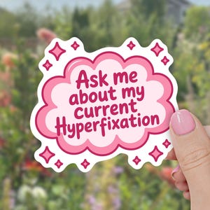 May include: A die-cut sticker shaped like a pink cloud with a white border. The sticker features the dark pink text "Ask me about my current Hyperfixation" in the center, surrounded by small, dark pink four-pointed star icons on the edges.