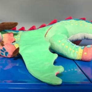 May include: A plush dragon toy featuring a light green body, large teal head with brown horns, and bright pink spikes running along its back. The creature has large green eyes, pink accents on its wings and feet, and a long, tapering tail.