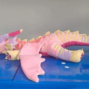 Puede incluir: Un peluche de dragón en tonos rosa, lavanda y crema. El dragón presenta grandes ojos azules, alas suaves y una cola larga con espinas amarillas a lo largo del lomo. El cuerpo tiene una textura estampada y los pies tienen huellas de color rosa.