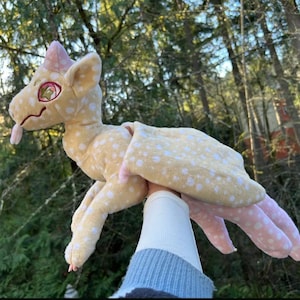 May include: A soft toy dragon, featuring a light yellow body with white polka dots. It has pink wings, feet, and a horn, along with a pink tongue. The dragon is held up against a blurred backdrop of trees.