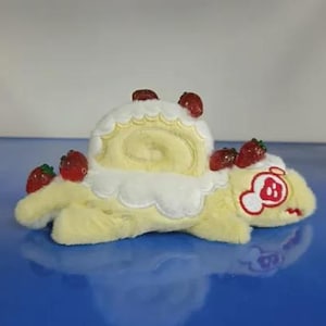 May include: A plush toy shaped like a yellow creature with a white cake roll on its back, topped with red strawberry accents. The creature has a red and white embroidered face on the side, featuring a closed eye and a small mouth.
