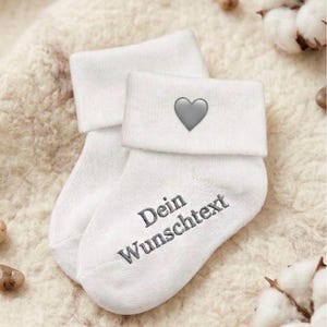 Könnte beinhalten: Ein Paar weiße Babysocken mit umgeschlagenem Bündchen und einem kleinen grauen Herz-Symbol. Auf dem Fußbereich der Socken ist der graue Text "Dein Wunschtext" in einer klaren, serifenlosen Schrift eingestickt, was auf ein personalisierbares Design hinweist.