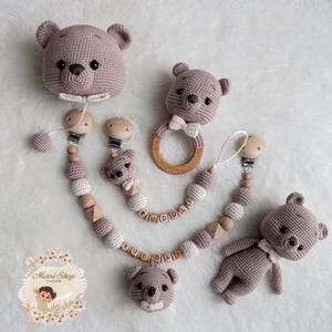 Può includere: Una collezione di accessori per neonati fatti a mano all'uncinetto nei toni malva, crema e legno. Il set include un porta ciuccio a forma di orso, un anello per la dentizione e un piccolo orsetto. Il porta ciuccio ha il nome "RUDOLF" scritto con perline di legno.