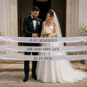 Könnte beinhalten: Ein weißes Satinband mit dem schwarz aufgedruckten Text JUST MARRIED, MR AND MRS JAN und 10.10.2026. Das Band wird horizontal gehalten und mit einer Schere durchtrennt, wobei es eine glatte, glänzende Oberfläche und klare Schriftzüge aufweist.