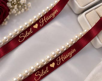 Può includere: Due nastri di raso bordeaux decorati con una fila di perle bianche. Ogni nastro presenta la scritta dorata Sibel ♥ Hüseyin in corsivo. I nastri sono posizionati vicino a scatole per anelli bianche e ad alcuni elementi floreali.