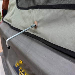 May include: A grey canvas vehicle tailgate cover with black trim and a central metal L-shaped rod protruding through a reinforced hole. Below the cover, a dark licence plate is visible with the yellow text WATER-WINTER WONDERLAND.