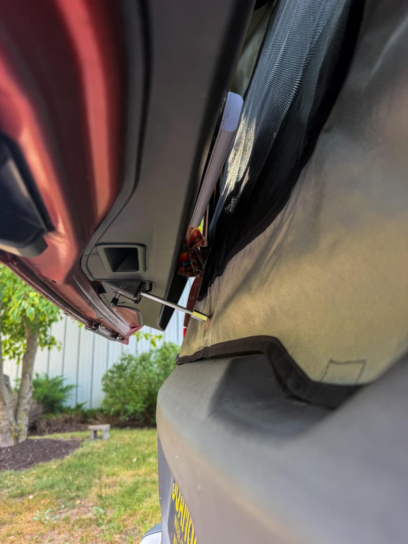 May include: A metal liftgate support strut attached to the interior frame of a vehicle's rear hatch. The silver-toned rod extends from a black plastic housing, connecting to a grey fabric panel that covers the rear window area of the car.