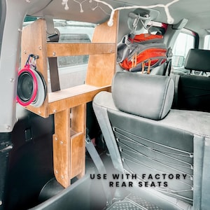 May include: A wooden shelf unit installed in the back of a car, with two shelves and a small storage compartment. The shelf unit is designed to be used with factory rear seats. A backpack hangs from a hook above the shelf unit.