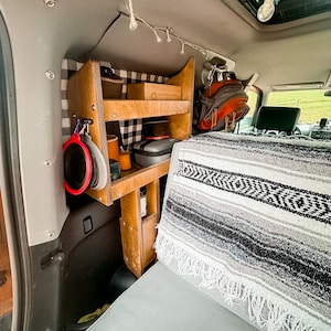 May include: A wooden shelf unit with two shelves is mounted in the back of a vehicle. The shelves are filled with various items, including a black and gray case, a brown box, and a few other items. A black and white blanket is draped over the back seat of the vehicle.
